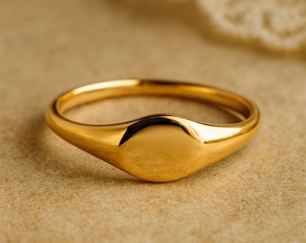 May include: A gold signet ring with a smooth, oval face. The ring is made of polished gold and has a simple, elegant design. The ring is shown on a neutral background.