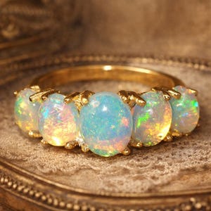 Könnte beinhalten: Ein goldener Ring mit fünf ovalen Opalen. Die Opale zeigen ein Farbspiel mit Blau-, Grün- und Gelbtönen. Der Ring liegt auf einem dekorativen, verzierten Tablett.