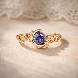 Può includere: Un anello color oro con una grande gemma rotonda e multicolore, incastonata in un design a mezzaluna. L'anello è adornato da delicati dettagli di foglie e viti e piccole gemme viola. L'anello è un gioiello.