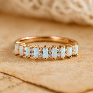 Op de afbeelding: Een gouden ring met een reeks rechthoekige opaalstenen en kleine diamanten. De opalen zijn melkachtig wit en de diamanten schitteren. De ring is gezet op een gouden band, wat een delicaat en elegant ontwerp creëert.