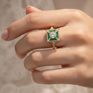 May include: A gold ring featuring a central round diamond surrounded by emerald-colored stones in a square design. The ring has a delicate band and is adorned with smaller diamonds. The ring is held up by a hand.