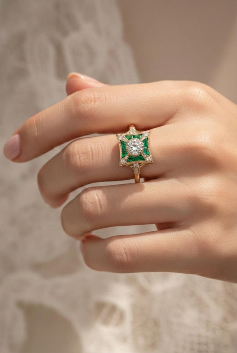 May include: A gold ring featuring a central round diamond surrounded by emerald-colored stones in a square design. The ring has a delicate band and is adorned with smaller diamonds. The ring is held up by a hand.