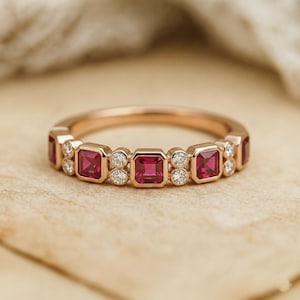 Könnte beinhalten: Ein roségoldener Bandring mit abwechselnd quadratisch geschliffenen Rubin-Edelsteinen und kleinen runden Diamanten. Die Rubine sind tiefrot und die Diamanten funkeln. Der Ring ist auf einem neutralen Hintergrund platziert.