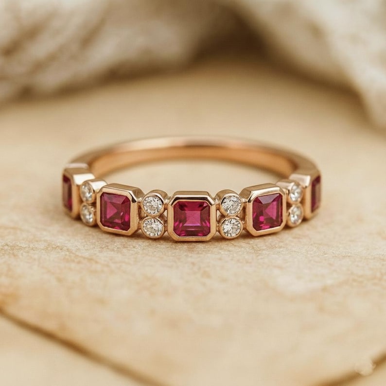Pode incluir: Um anel de banda em ouro rosa com altern&acirc;ncia de pedras preciosas de rubi de corte quadrado e pequenos diamantes redondos. Os rubis s&atilde;o de cor vermelho profundo e os diamantes brilham. O anel est&aacute; sobre um fundo neutro.