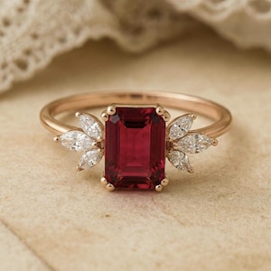 May include: A rose gold ring with a large, rectangular, deep red gemstone. Either side of the central stone are clusters of marquise-cut, clear gemstones. The ring has a slender, polished metal band.
