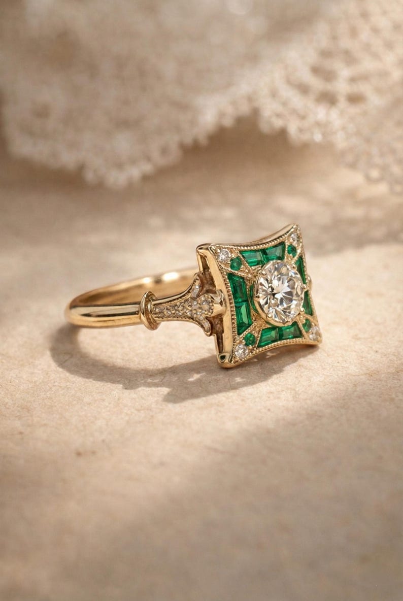 May include: A gold ring with a square emerald and diamond cluster design. The center features a round diamond, surrounded by emeralds and smaller diamonds. The ring has a detailed band and setting, showcasing a vintage or Art Deco style.