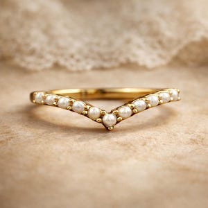 Puede incluir: Un anillo dorado con un diseño en forma de V. El anillo está adornado con pequeñas perlas blancas redondas. El anillo se encuentra sobre un fondo neutro, resaltando el elegante diseño de la joya. El anillo es una pieza de joyería delicada.