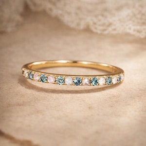 Op de afbeelding: Een gouden ring met een rij kleine, ronde edelstenen. De edelstenen wisselen af tussen lichtblauw, roze en helder. De ring is geplaatst tegen een neutrale achtergrond.