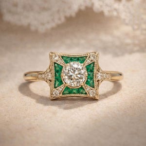 May include: A gold ring featuring a central round diamond, surrounded by emeralds and smaller diamonds in a square design. The ring has a vintage, art deco style with intricate detailing.