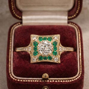 May include: A gold ring with a large, round, clear center stone, surrounded by emerald-green stones and smaller clear stones. The ring is set in a square design and is displayed in a burgundy ring box.