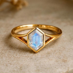 Op de afbeelding: Een gouden ring met een zeshoekige, lichtblauwe edelsteen. De band van de ring is van gepolijst goud, met een geometrisch ontwerp dat de edelsteen ondersteunt. De edelsteen heeft een subtiele, iriserende kwaliteit.