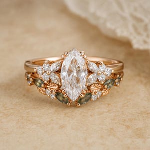 May include: A rose gold engagement ring set with a marquise-cut diamond centre stone. The band is decorated with floral and leaf accents, and small accent diamonds. The set includes a matching wedding band with similar floral details.