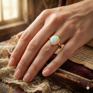 May include: A gold ring featuring an oval opal gemstone. The opal displays a play of color with iridescent hues. The ring is set in a simple gold band. The hand is resting on a vintage-style surface.
