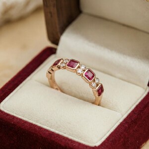 Pode incluir: Um anel de ouro com pedras vermelhas quadradas alternadas e pequenas pedras claras redondas. O anel &eacute; apresentado numa caixa de joias de veludo vermelho com interior creme. O anel &eacute; uma joia.