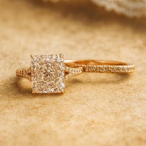 Puede incluir: Un anillo de compromiso de oro con un diamante de talla radiante y una alianza a juego. El anillo de compromiso presenta un gran diamante de talla cuadrada y diamantes más pequeños en la banda y los hombros. La alianza también está adornada con pequeños diamantes.