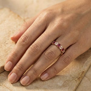 Pode incluir: Um anel de ouro com pedras preciosas vermelhas e claras alternadas. O anel apresenta pedras vermelhas retangulares e pedras claras redondas embutidas em uma faixa de ouro. O anel &eacute; usado em um dedo.