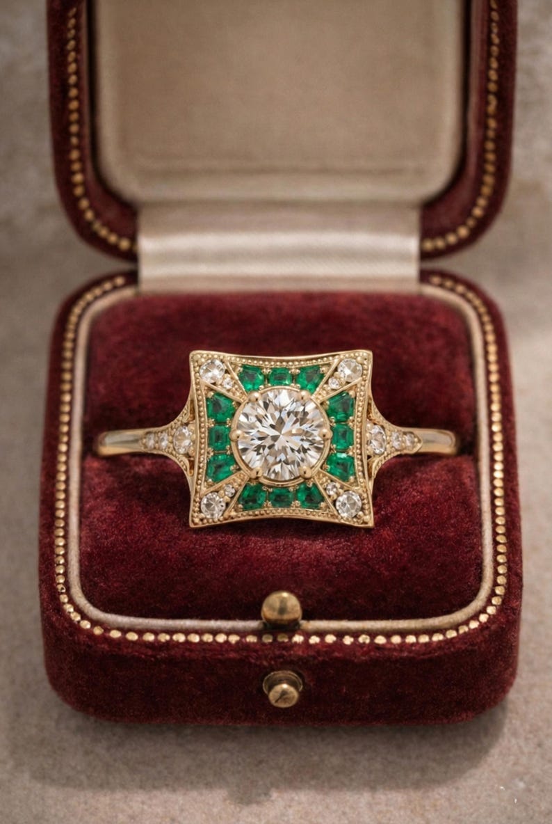 May include: A gold ring with a large, round, clear center stone, surrounded by emerald-green stones and smaller clear stones. The ring is set in a square design and is displayed in a burgundy ring box.