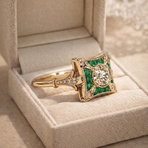 May include: A gold ring with a square emerald and diamond setting, displayed in a beige ring box. The ring features a central round diamond surrounded by emeralds and smaller diamonds. The band is also adorned with diamonds.