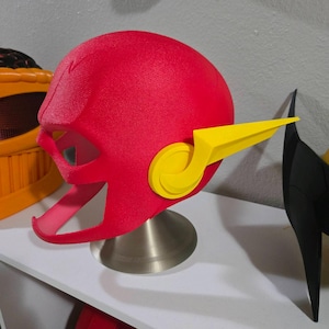 May include: A red superhero helmet with yellow side wings, displayed on a silver stand. The helmet has a textured surface and a distinctive design, reminiscent of a comic book character. The helmet is a vibrant red color.