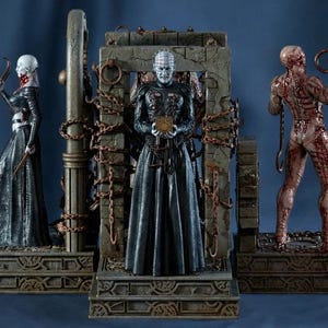 May include: Three detailed figurines from the horror film franchise, Hellraiser. The central figure, Pinhead, stands in front of a stone structure. Other figures include a female Cenobite and a flayed Cenobite, all on decorative bases.