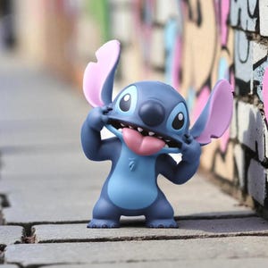 K&ouml;nnte beinhalten: Eine blaue und hellblaue Stitch-Figur mit rosa Ohren und Zunge, die ein albernes Gesicht macht. Die Figur steht auf einer grauen Ziegeloberfl&auml;che, mit einer bunten Graffiti-Wand im Hintergrund.