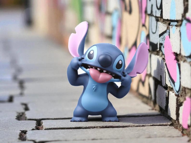 K&ouml;nnte beinhalten: Eine blaue und hellblaue Stitch-Figur mit rosa Ohren und Zunge, die ein albernes Gesicht macht. Die Figur steht auf einer grauen Ziegeloberfl&auml;che, mit einer bunten Graffiti-Wand im Hintergrund.