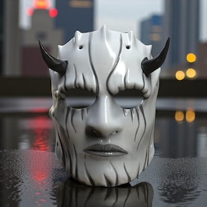May include: A white ceramic mask with black horns and black vertical lines. The mask has eye holes, a nose, and a mouth. The mask is on a reflective surface with a blurred city background.