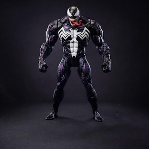 May include: A Venom action figure in a menacing pose. The figure is mainly black with purple and white details. A white spider emblem is on the chest. The figure's mouth is open, showing a red tongue. The background is dark. The figure is approximately 18 cm tall.