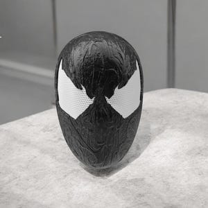 Peut inclure: Un masque noir et blanc avec une surface texturée, présentant de grandes formes d'yeux blancs. Le masque est une réplique du masque d'un personnage de bande dessinée. Le masque est exposé sur une surface de couleur claire.