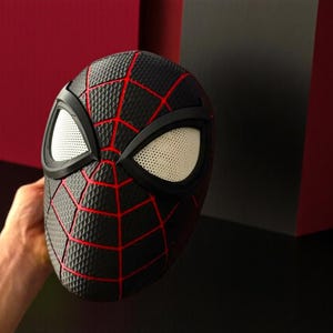 May include: Black Spider-Man mask with red web lines and white eye lenses. The mask has a textured surface and is held by a hand. The mask is a superhero costume accessory.