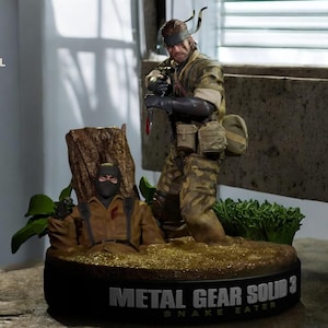 Könnte beinhalten: Detaillierte Figur aus Metal Gear Solid 3: Snake Eater. Die Figur zeigt zwei Charaktere in Militäruniform, einer hält ein Messer. Der Sockel trägt den Titel des Spiels. Die Figur steht auf einem detaillierten Sockel mit Blättern.