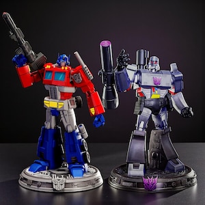 May include: Two action figures of Transformers characters Optimus Prime and Megatron. Optimus Prime is red, blue, and gray, holding a black weapon. Megatron is gray and purple, holding a black weapon. Both figures stand on circular bases.