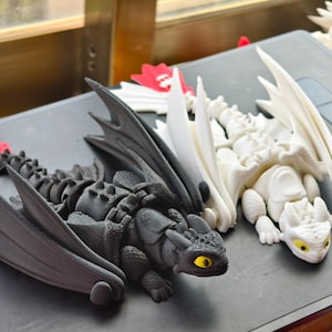 Puede incluir: Tres figuras de dragones impresas en 3D. Una es negra con alas grises y una aleta caudal roja, la otra es blanca, y la tercera es una aleta caudal de dragón roja y blanca. Los dragones están sobre una superficie gris.