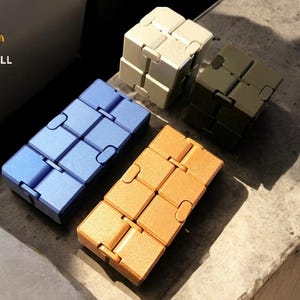 May include: A selection of fidget toys in shades of blue, orange, cream, and olive green. The toys are composed of interconnected cubes, designed for manipulation. The image is well-lit, highlighting the colours and textures of the toys.