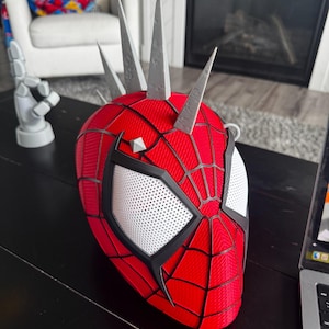 May include: A red and black Spider-Man mask with white eye lenses and gray spikes on top. The mask has a web pattern and is sitting on a black surface.