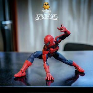 K&ouml;nnte beinhalten: Eine Spider-Man-Actionfigur in dynamischer Pose, gekleidet in einem rot-blauen Anzug. Die Figur ist mit Netzmustern detailliert und vor einem verschwommenen Hintergrund platziert. Der Text "PATULL SPIDER-MAN" ist sichtbar.