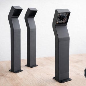 Könnte beinhalten: Drei schwarze Sicherheitskameras im modernen Stil mit einem schlanken, eckigen Design. Eine Kamera zeigt die internen Komponenten. Die Kameras sind auf einer Holzoberfläche montiert. Das Bild enthält einen "Made in Germany" Aufkleber.