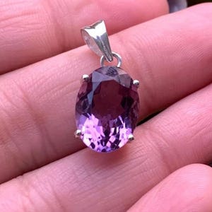 Puede incluir: Un colgante de plata con una amatista ovalada de color p&uacute;rpura oscuro. La piedra preciosa est&aacute; engastada en un bisel de plata y tiene un acabado pulido. El colgante est&aacute; suspendido de una peque&ntilde;a fianza de plata. Fondo borroso, con follaje verde.