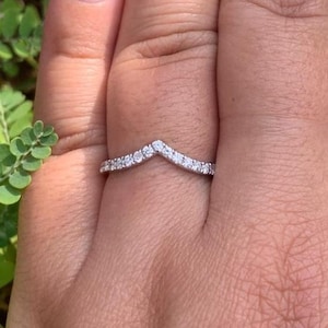May include: A silver-toned chevron-shaped ring featuring a row of small, clear, round-cut stones set along the band. The ring has a pointed V-shape design and a polished metallic finish, displaying a delicate and symmetrical appearance on the finger.
