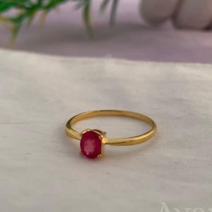 Può includere: Un anello in oro con una singola pietra preziosa rossa di forma ovale incastonata al centro. La fascia è sottile e lucida, con una finitura metallica liscia. La parola Avanira è visibile sullo sfondo, parzialmente oscurata da fogliame verde.