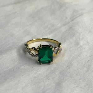 Peut inclure: Une bague dorée avec une pierre précieuse vert émeraude rectangulaire au centre. La monture présente un motif décoratif avec de petites pierres claires de chaque côté de l'émeraude. La bague est posée sur un fond blanc.