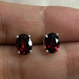 Pode incluir: Um par de brincos de tarracha com granadas vermelhas ovais cravadas em garras de prata. As pedras apresentam um corte facetado que reflete a luz, e a palavra Avanira é visível no fundo.