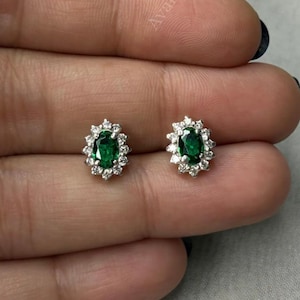 May include: A pair of stud earrings featuring oval-cut emeralds surrounded by a halo of small, clear gemstones. The earrings are set in a silver-colored metal and are displayed on a hand.