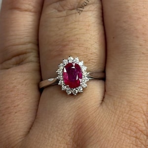 Puede incluir: Un anillo de tono plateado con una piedra preciosa de color rojo intenso de corte ovalado en el centro, rodeada por un halo de pequeñas piedras claras de corte redondo. La palabra Avanira es visible al fondo. El anillo tiene una banda de metal pulido.