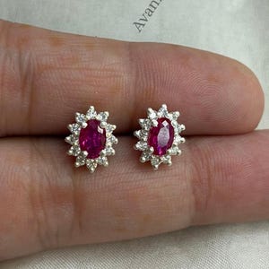 Puede incluir: Un par de pendientes de botón con piedras centrales de color rosa intenso en forma ovalada, rodeadas por un halo de pequeñas piedras blancas transparentes engastadas en un marco metálico. La palabra Avanira aparece impresa en negro.