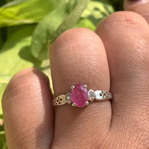 Op de afbeelding: Een zilveren ring met een ovaalvormige, helder roze edelsteen. De ring is versierd met kleine, heldere stenen aan weerszijden van de hoofdsteen en een decoratieve band. De ring wordt getoond op een hand, tegen een achtergrond van groene bladeren.