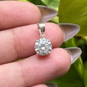 Puede incluir: Un colgante de tono plateado con una piedra preciosa redonda, transparente y facetada, sujeta en un engaste de cuatro garras con una pequeña anilla en la parte superior. La palabra Avanira está impresa en letras negras pequeñas en el lateral y abajo.