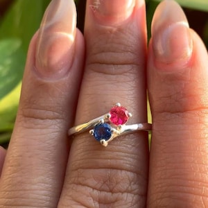 May include: A silver ring featuring a blue and a red gemstone. The ring's band curves gracefully, with the gemstones set close together. The ring is shown on a hand, with the word "Avanina" visible in the background.