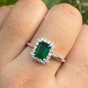 Peut inclure: Une bague argentée ornée d'une pierre précieuse verte rectangulaire taille émeraude au centre, entourée d'un halo de petites pierres claires taille ronde. L'anneau est fin et métallique, et le mot Avanira est visible sur l'image.