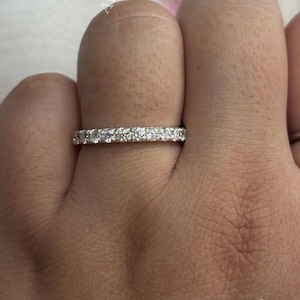 Könnte beinhalten: Ein silberfarbener Eternity-Ring mit einer durchgehenden Reihe kleiner, klarer, rund geschliffener Steine, die entlang des gesamten sichtbaren Umfangs des Rings gefasst sind. Das Wort Avanira ist in weißer Schrift in den Ecken zu sehen.